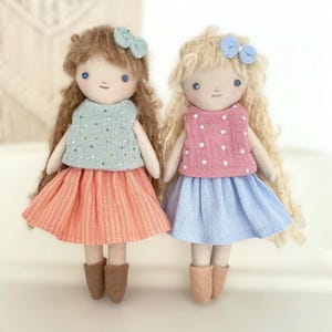 Peut inclure: Deux poupées en tissu faites à la main. L'une a des cheveux bruns, un nœud bleu, un haut bleu clair à pois blancs et une jupe orange. L'autre a des cheveux blonds, un nœud bleu, un haut rose à pois blancs et une jupe bleue.