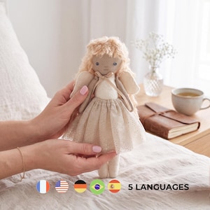 Op de afbeelding: Een stoffen pop met lang, golvend blond haar, gekleed in een crèmekleurig lijfje en een glitterende, lichtbeige tule rok. De pop heeft kleine, donker geborduurde ogen, een eenvoudige gezichtsuitdrukking en kleine vleugels op de rug.
