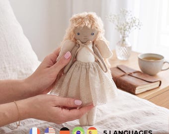 May include: A handmade fabric doll with long, wavy blonde yarn hair, wearing a cream-colored bodice and a shimmering, glittery tulle skirt. The doll features simple embroidered facial details, small fabric wings on the back, and a delicate ribbon tie at the neck.