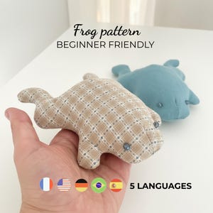 May include: Two handmade frog plushies. One frog is tan with a plaid pattern, and the other is light blue. The text "Frog pattern BEGINNER FRIENDLY" is at the top of the image. The image also includes the flags of five different languages.