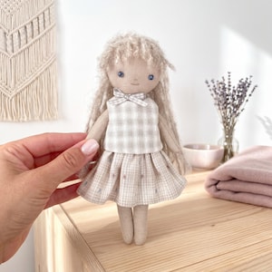 Pode incluir: Uma boneca de tecido feita à mão com cabelo comprido de lã bege texturizada e olhos azuis bordados. A boneca usa um top sem mangas e uma saia plissada, ambos com um padrão xadrez bege claro e branco com um pequeno laço no decote.