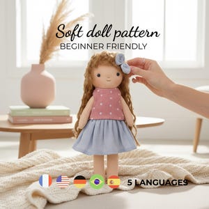 May include: A soft doll with long brown hair, wearing a pink polka-dot top and a blue skirt. The doll has a blue bow and is labeled "Soft doll pattern BEGINNER FRIENDLY". The image includes flags representing 5 languages.