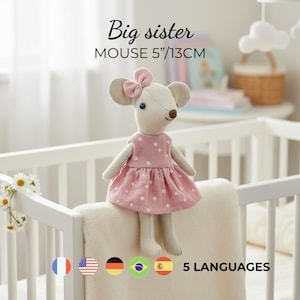 Pode incluir: Um brinquedo de rato de pelúcia, com 13 cm de altura, vestindo um vestido rosa com bolinhas e um laço combinando. O texto "Big sister" e "MOUSE 5"/13CM" está acima do brinquedo. O rato está em um berço branco.
