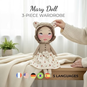 May include: A Mary Doll with a 3-piece wardrobe. The doll wears a brown bear-eared hat, a tan sweater, and a floral dress. The doll is on a wooden surface with a white blanket. The image includes the text "Mary Doll 3-PIECE WARDROBE" and flags for 5 languages.