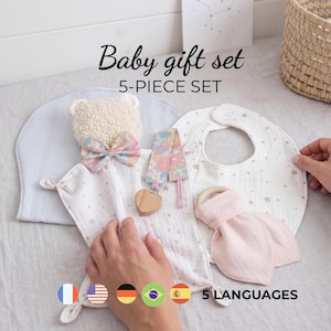 Puede incluir: Un set de regalo para bebé de 5 piezas que incluye una manta de seguridad de oso de peluche color crema con lazo floral, un clip para chupete floral con corazón de madera, un babero blanco con estrellas doradas, uno azul claro y un mordedor de tela.