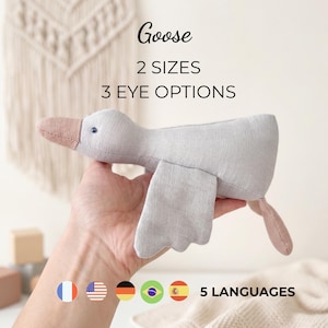Może przedstawiać: Jasnoszara pluszowa gęś z beżowym dziobem i ogonem, posiadająca małe ciemne oko. Widoczny jest tekst Goose, 2 SIZES, 3 EYE OPTIONS oraz 5 LANGUAGES, a także pięć małych okrągłych ikon flag reprezentujących różne kraje na świecie.