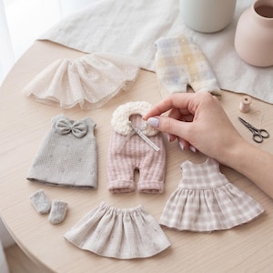 May include: A collection of miniature doll clothing including a beige tulle skirt, a grey gingham dress with a bow, a pink jumpsuit with a white faux-fur collar, a beige plaid dress, a small beige skirt, and two tiny grey socks arranged on a table.