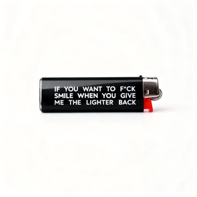 Pu&ograve; includere: Un accendino nero con la scritta "IF YOU WANT TO F*CK SMILE WHEN YOU GIVE ME THE LIGHTER BACK" in bianco. L'accendino ha una parte superiore in metallo argentato e una leva in plastica rossa.