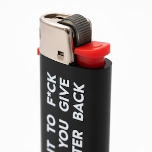 Pu&ograve; includere: Accendino nero con la scritta bianca "I WANT TO F*CK YOU GIVE TER BACK". Presenta una parte superiore in metallo argentato e un pulsante di accensione rosso. Sfondo bianco.