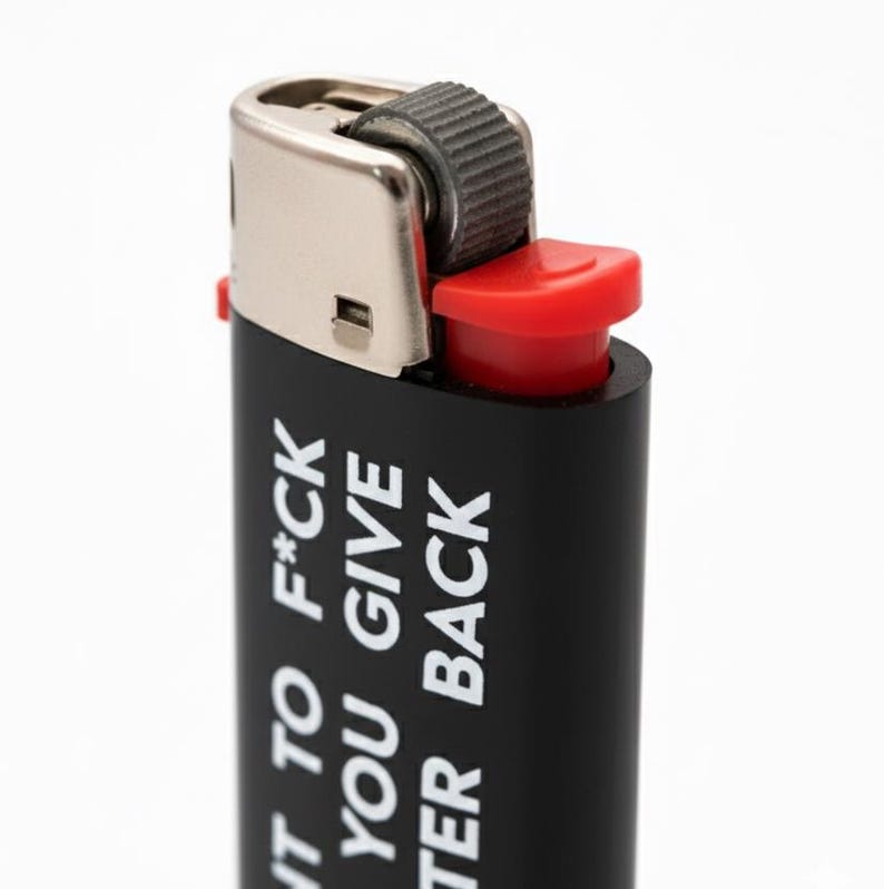 Pu&ograve; includere: Accendino nero con la scritta bianca "I WANT TO F*CK YOU GIVE TER BACK". Presenta una parte superiore in metallo argentato e un pulsante di accensione rosso. Sfondo bianco.