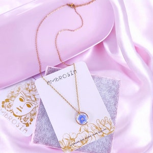 May include: A gold chain necklace with a small, round pendant featuring a pressed blue flower. The pendant is on a white card with the text "AMBROSIÄ" and "FLOWERS ARE MORE THAN BEAUTY".