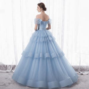 May include: A light blue off-the-shoulder gown with a layered skirt. The dress features a corset back and a flowing, tiered design. The gown is made of a shimmering fabric, perfect for formal events.