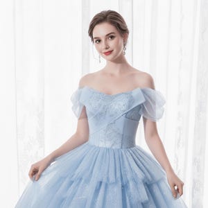 May include: A light blue off-the-shoulder gown with a layered skirt. The bodice features intricate embroidery and a fitted waist. The dress has sheer, ruffled sleeves and a flowing, tiered skirt, creating an elegant silhouette.