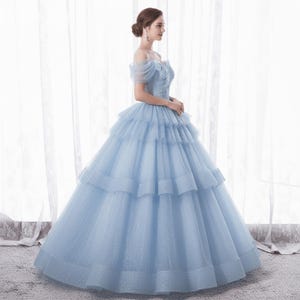 May include: A light blue formal gown with an off-the-shoulder neckline and layered skirt. The bodice features delicate embroidery, and the dress has a full, flowing silhouette. The gown is made of a shimmering fabric, perfect for special occasions.