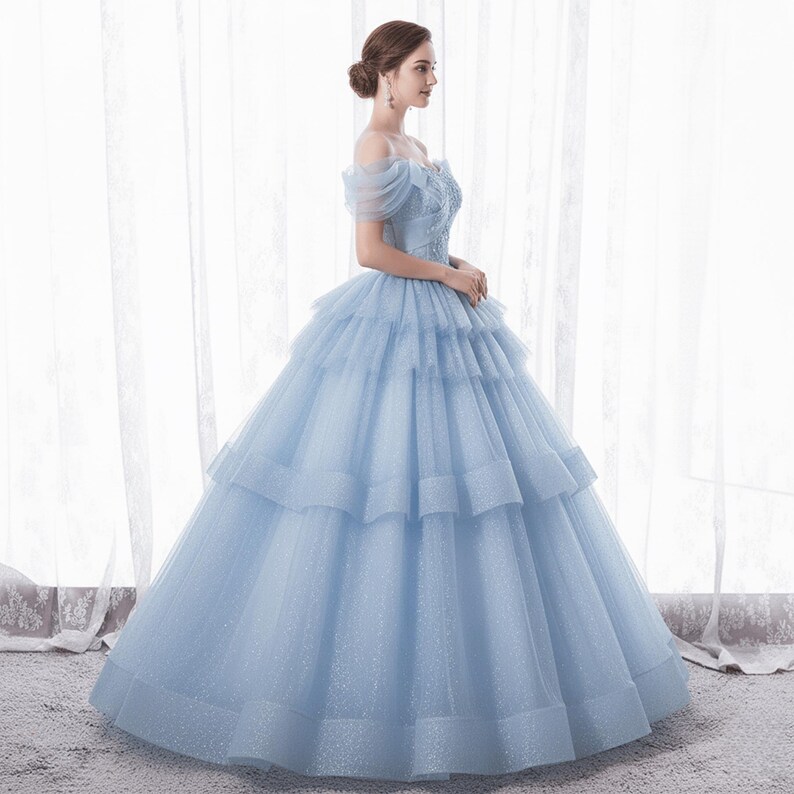 May include: A light blue formal gown with an off-the-shoulder neckline and layered skirt. The bodice features delicate embroidery, and the dress has a full, flowing silhouette. The gown is made of a shimmering fabric, perfect for special occasions.