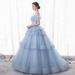 May include: A light blue off-the-shoulder gown with a layered skirt. The dress features a fitted bodice and a full, flowing skirt. The fabric has a subtle shimmer, and the gown is floor-length.
