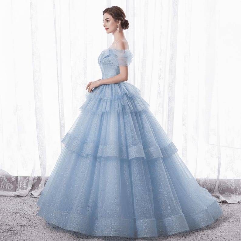 May include: A light blue off-the-shoulder gown with a layered skirt. The dress features a fitted bodice and a full, flowing skirt. The fabric has a subtle shimmer, and the gown is floor-length.