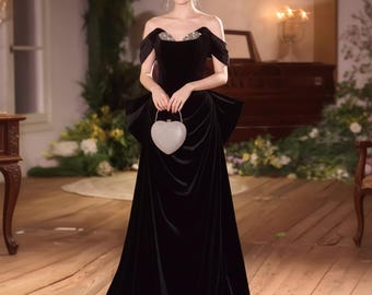 May include: A black off-the-shoulder velvet gown with a sweetheart neckline, embellished with sparkling details. The dress features a large bow detail at the waist and is paired with a heart-shaped, silver clutch. The gown is floor-length.