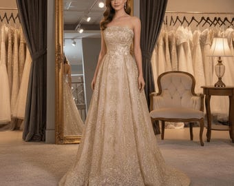 Puede incluir: Un vestido de novia sin tirantes, largo hasta el suelo, color champán, con intrincados bordados dorados. El vestido presenta una falda amplia y se exhibe en una tienda de novias, con otros vestidos visibles en el fondo.