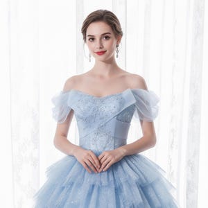 May include: A light blue off-the-shoulder gown with a layered tulle skirt. The bodice features intricate beadwork and a sweetheart neckline. The dress has sheer, puffed sleeves and is suitable for formal events.