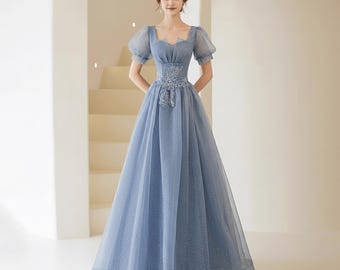 Könnte beinhalten: Ein hellblaues Abendkleid mit einem figurbetonten Oberteil und einem weiten Rock. Das Kleid hat kurze, gepuffte Ärmel und zarte Blumenstickereien an der Taille. Der Stoff schimmert leicht, und der Ausschnitt ist quadratisch.