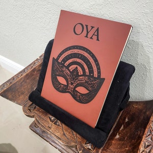 May include: A reddish-brown journal with the word "OYA" and a black mask design. The journal is propped up on a black pillow, resting on a carved wooden stand. The mask design includes a rainbow and intricate details.