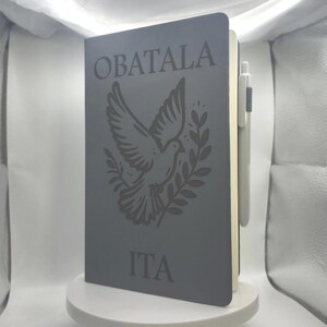 May include: Gray notebook with the word "OBATALA" at the top and "ITA" at the bottom. A dove with olive branches is in the center. A gray pen is attached to the side of the notebook.