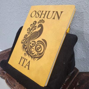 May include: A mustard-colored journal with the word "OSHUN" and "ITA" engraved on the cover. A detailed peacock design is etched below "OSHUN". The journal is propped up on a black surface.