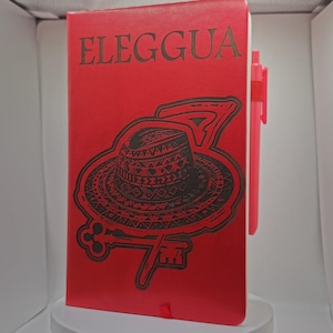 May include: Red notebook with the word "ELEGGUA" in dark letters. The cover features a black and brown graphic of a hat, key, and number seven. A pink pen is attached to the side of the notebook.