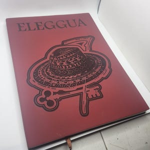 May include: A red notebook with the word "ELEGGUA" printed at the top. The cover features a detailed illustration of a hat, key, and decorative border in a darker shade of red. A brown ribbon bookmark is visible.