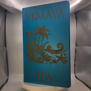 May include: A teal-colored notebook with the word "YEMAYA" at the top and "ITA" at the bottom. The notebook features a gold-colored illustration of palm trees, seashells, a sailboat, and a starfish. The notebook is standing upright.
