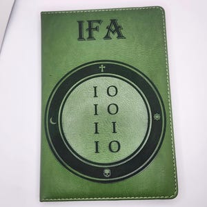 May include: Green leather-like journal with the letters "IFA" at the top. A circular design in the center features the letters "IO" repeated, with a cross, skull, and other symbols. The journal has a stitched border.