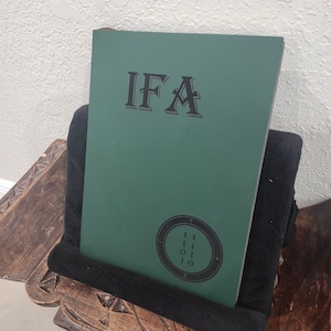 May include: A green book with the letters "IFA" in black, resting on a black cushioned stand. Below the letters is a circular design with the numbers "111010". The stand is on a wooden surface.