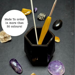 May include: A black hexagonal container holding two crochet hooks, one white and one yellow. The container is surrounded by various polished gemstones in shades of purple, gold, and black. Text in a white circle reads "Made To order in more than 50 colours!"