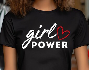 以下が含まれることがあります： 白い筆記体で「girl POWER」と赤いハートのグラフィックがプリントされた黒いTシャツ。レンガの壁と黒い風船の前で、人がそのシャツを着ています。
