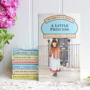 May include: A stack of five paperback children's books with titles like "Fairy Tales", "What Katy Did", "Alice in Wonderland", "Heidi", "The Secret Garden", and "Black Beauty". The books are stacked on a white lace tablecloth. A blue and white book titled "A Little Princess" by Frances Hodgson Burnett is in the background.