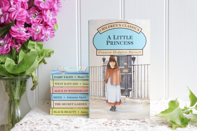 May include: A stack of five paperback children's books with titles like "Fairy Tales", "What Katy Did", "Alice in Wonderland", "Heidi", "The Secret Garden", and "Black Beauty". The books are stacked on a white lace tablecloth. A blue and white book titled "A Little Princess" by Frances Hodgson Burnett is in the background.