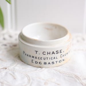 May include: A small, off-white ceramic jar with a rounded shape. The jar has a slightly aged appearance with faded text that reads "T. CHASE, PHARMACEUTICAL CHEMIST, EDG BASTON." The jar sits on a white, textured surface.