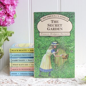 May include: A stack of classic children's books with colorful covers, including "Black Beauty", "A Little Princess", "Fairy Tales", "What Katy Did", "Alice in Wonderland", and "Heidi". The top book is "The Secret Garden" by Frances Hodgson Burnett, featuring a girl in a white dress and a straw hat opening a gate to a lush green garden.