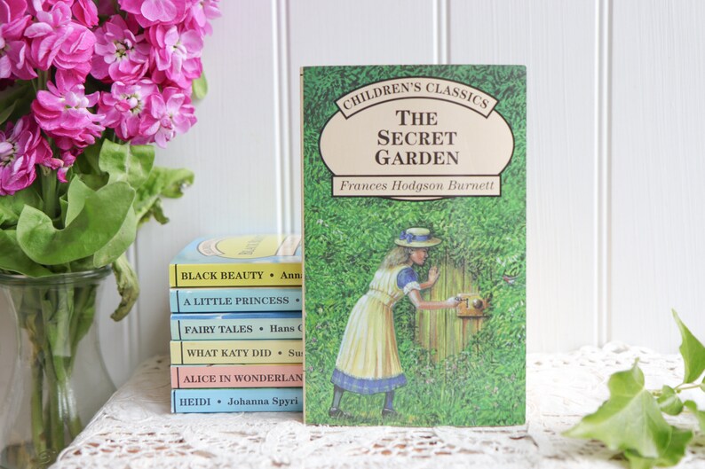 May include: A stack of classic children's books with colorful covers, including "Black Beauty", "A Little Princess", "Fairy Tales", "What Katy Did", "Alice in Wonderland", and "Heidi". The top book is "The Secret Garden" by Frances Hodgson Burnett, featuring a girl in a white dress and a straw hat opening a gate to a lush green garden.