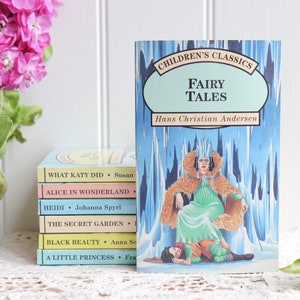 May include: A stack of five paperback books with colorful covers featuring titles like "What Katy Did", "Alice in Wonderland", "Heidi", "The Secret Garden", "Black Beauty", and "A Little Princess".  The top book is a blue and green paperback titled "Children's Classics Fairy Tales" by Hans Christian Andersen.  The book cover features an illustration of a woman in a green dress and fur cloak sitting on a throne.