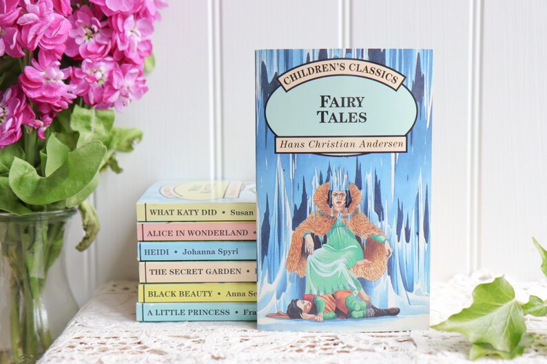 May include: A stack of five paperback books with colorful covers featuring titles like "What Katy Did", "Alice in Wonderland", "Heidi", "The Secret Garden", "Black Beauty", and "A Little Princess".  The top book is a blue and green paperback titled "Children's Classics Fairy Tales" by Hans Christian Andersen.  The book cover features an illustration of a woman in a green dress and fur cloak sitting on a throne.