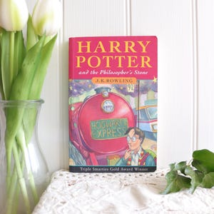 May include: A hardback book titled Harry Potter and the Philosopher's Stone by J.K. Rowling. The cover features a large red steam locomotive labelled Hogwarts Express, a character with glasses, and the text Triple Smarties Gold Award Winner.