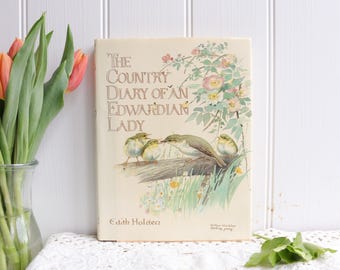 May include: A vintage book titled "The Country Diary of an Edwardian Lady" with a floral and bird illustration on the cover. The book is cream-colored and sits on a white lace tablecloth. A vase of orange tulips is to the left.