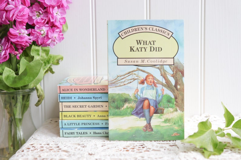 May include: A stack of classic children's books with titles like "Alice in Wonderland", "Heidi", "The Secret Garden", "Black Beauty", "A Little Princess", and "Fairy Tales".  The top book is "What Katy Did" by Susan M. Coolidge, featuring an illustration of a girl on a swing.