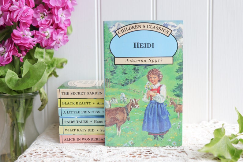 May include: A stack of vintage children's books with colorful covers. The top book is titled "Heidi" by Johanna Spyri. The book features a girl in a blue dress holding flowers and two goats in a green field.