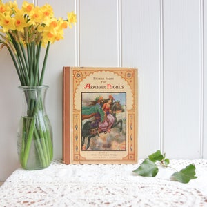 May include: A vintage book titled "Stories from the Arabian Nights" with a colorful illustration of a man and woman riding horses on a cloud. The book is on a white lace tablecloth with a vase of yellow daffodils in the background.