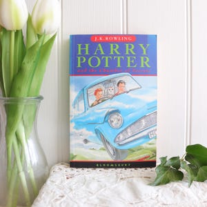May include: A hardcover book titled Harry Potter and the Chamber of Secrets by J.K. Rowling. The cover features a blue flying car with two people and an owl inside, set against a sky background. The publisher Bloomsbury is printed at the bottom.