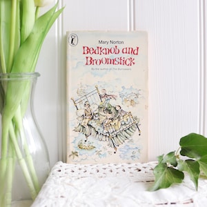 May include: A paperback book titled Bedknob and Broomstick by Mary Norton, featuring a whimsical illustration of children on a flying bed. The cover has a cream background with red text and a small penguin logo at the top left corner.
