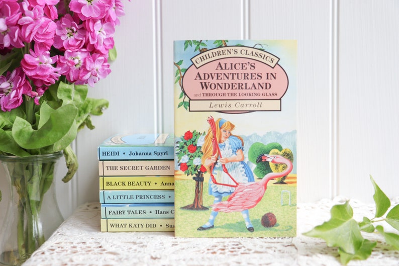May include: A stack of classic children's books with colorful covers, including "Heidi", "The Secret Garden", "Black Beauty", "A Little Princess", "Fairy Tales", and "What Katy Did".  The top book is "Alice's Adventures in Wonderland and Through the Looking Glass" by Lewis Carroll, featuring an illustration of Alice playing croquet with a flamingo.